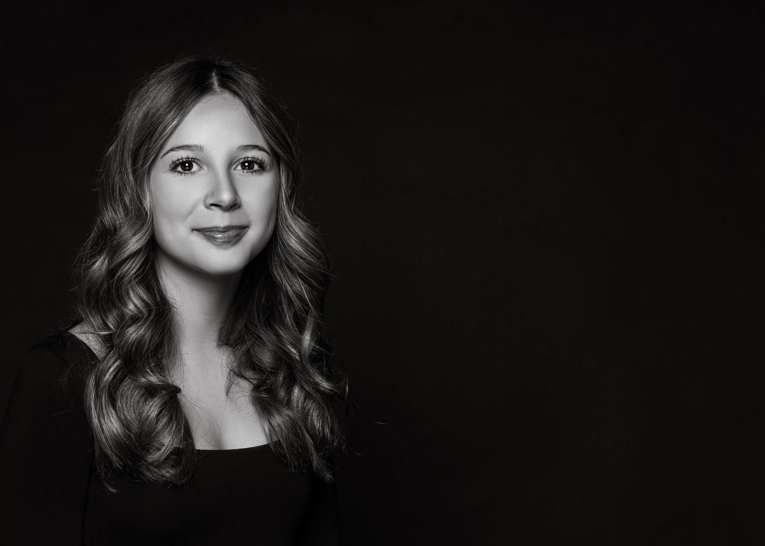 Black and white studio portrait featuring wavy hair and gentle smile against dark background.
