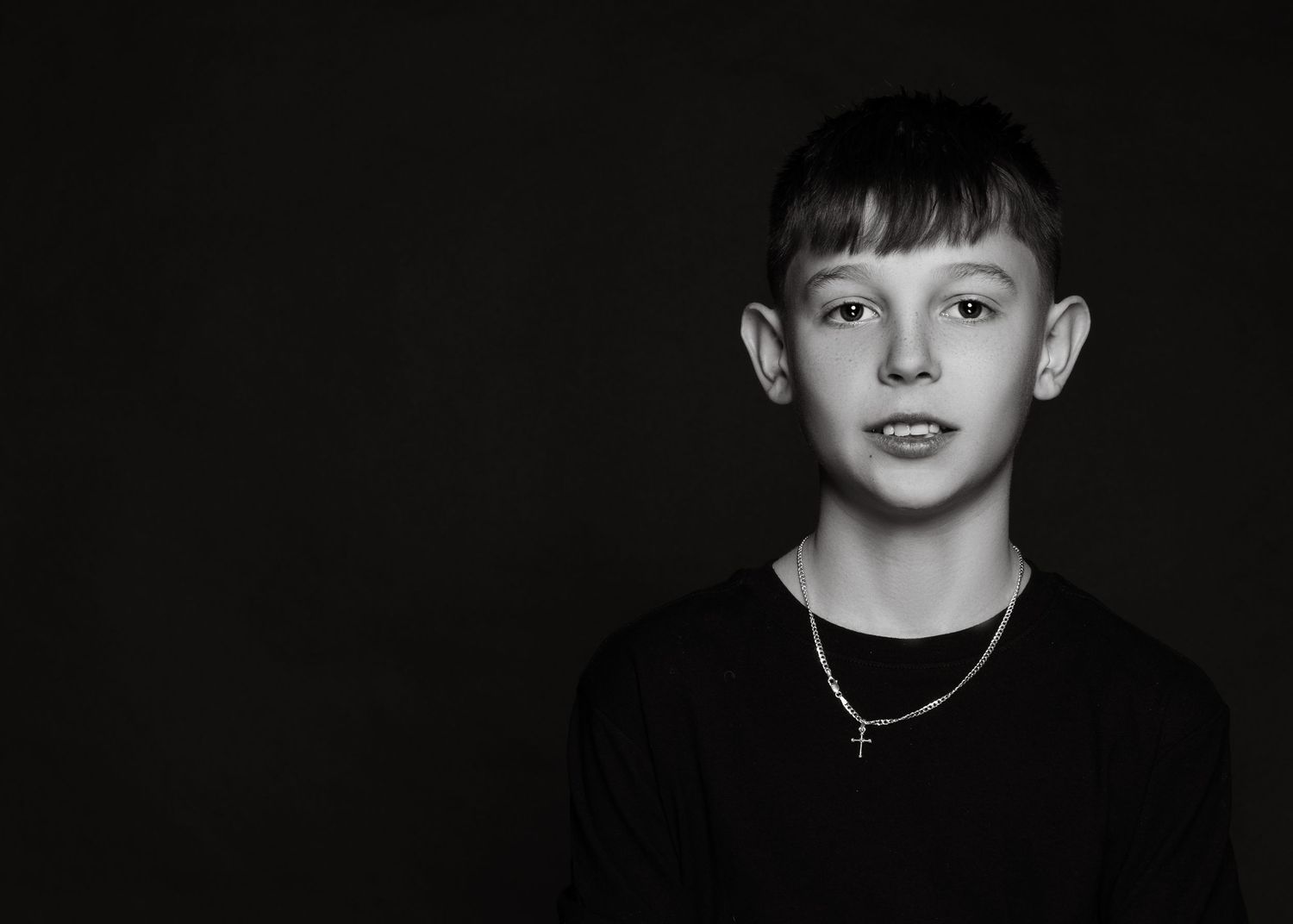 Black and white studio portrait of a young person with dark hair wearing a simple necklace against a dark background.