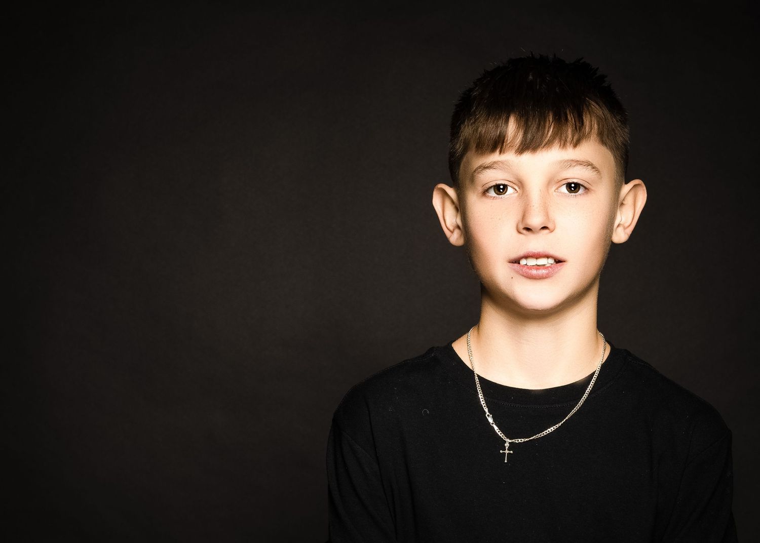 Head and shoulders portrait of young person wearing black top and necklace against dark background.