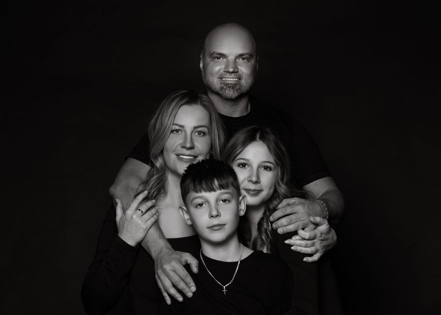 A warm black and white family portrait showing a close-knit group of four embracing against a dark background.