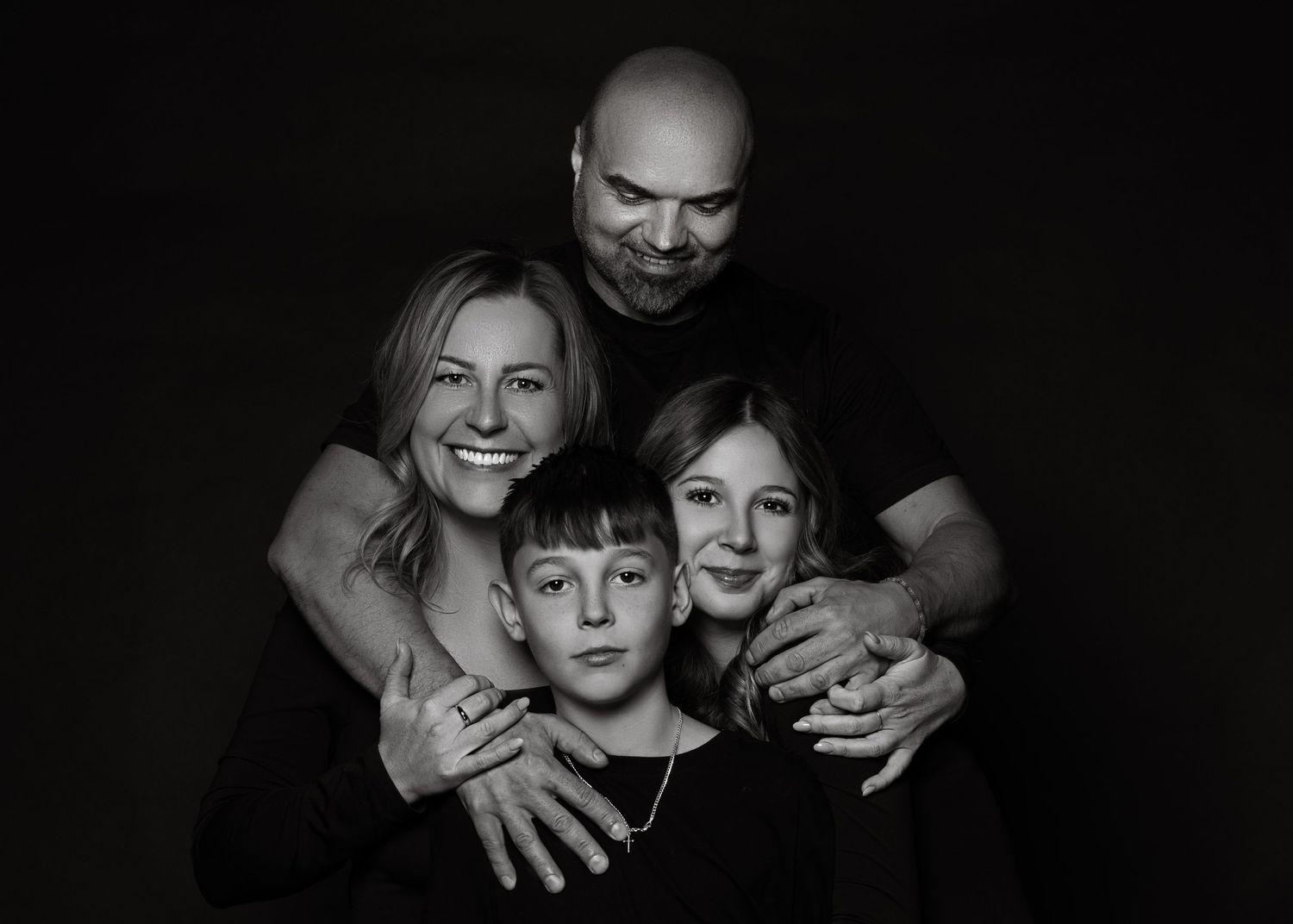 Black and white family portrait with dramatic lighting showing warm embrace against dark background.