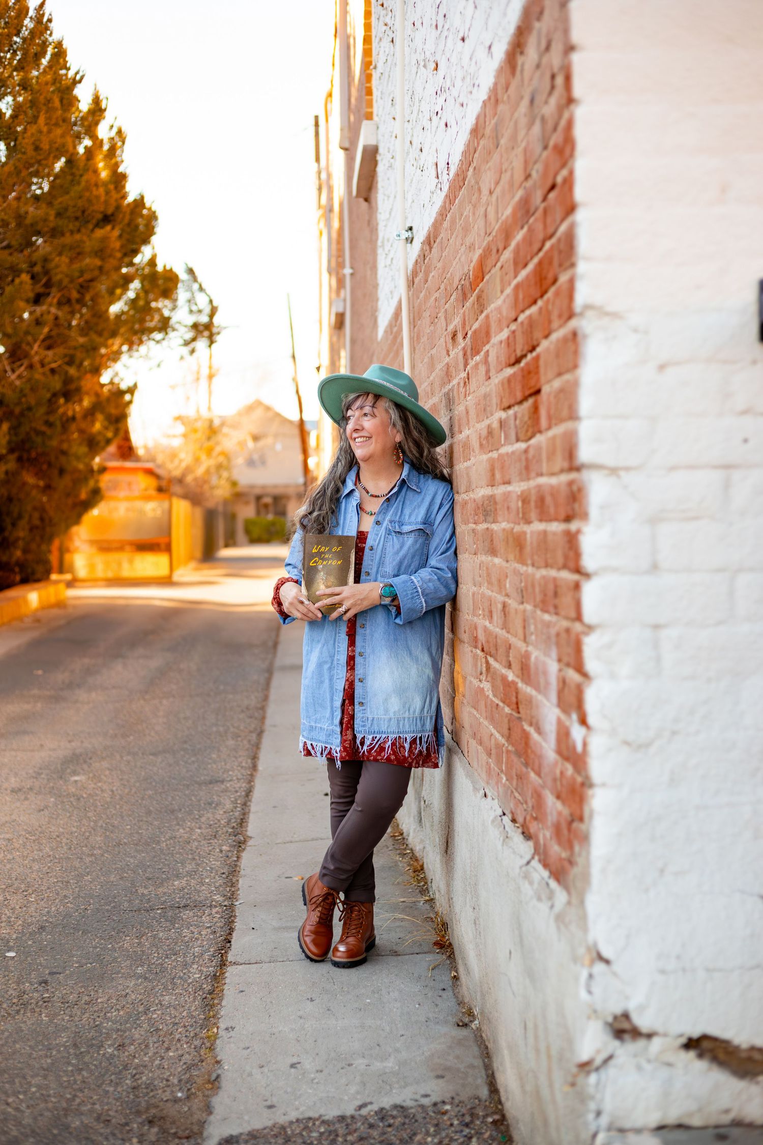 Casual outdoor photoshoot against brick wall at sunset with denim jacket and teal hat.