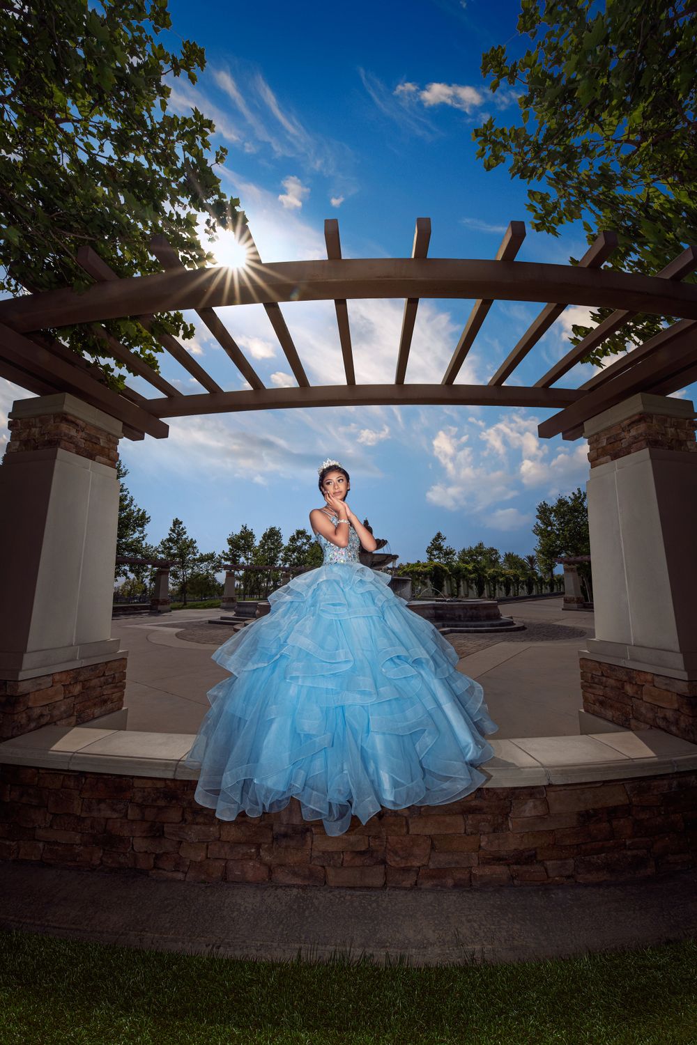 A stunning blue quinceañera dress glows in the sunlight under an elegant garden pergola with stone pillars.