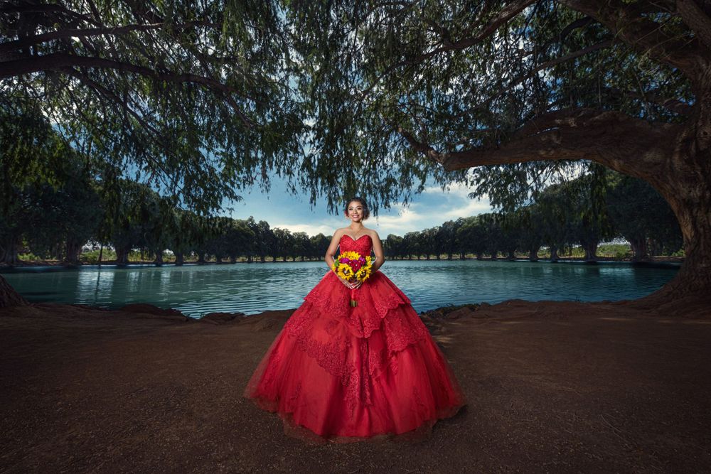A vibrant red ball gown stands out against a serene lakeside backdrop under hanging tree branches.