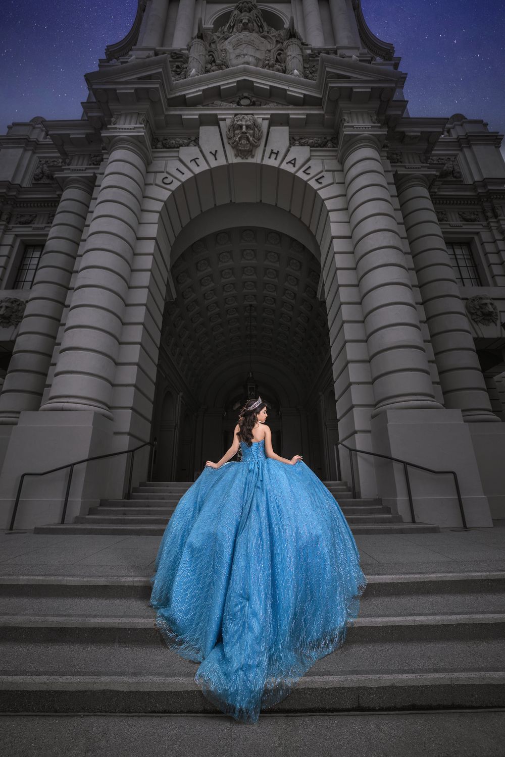 A figure in a flowing blue ballgown stands on stone steps of a grand classical building at dusk.