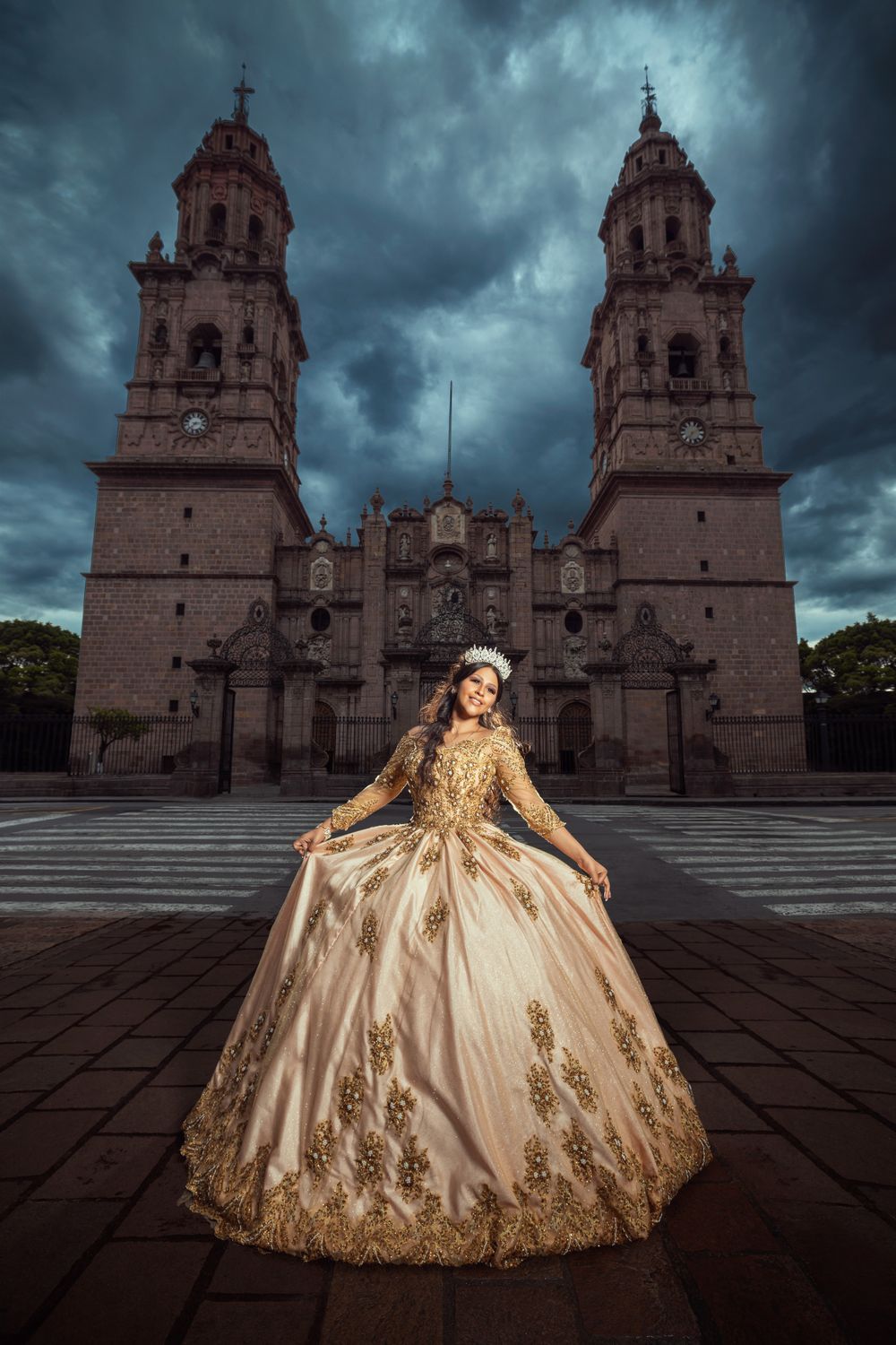A regal quinceanera dress glows in golden embroidery against the dramatic backdrop of a Mexican cathedral under stormy skies.