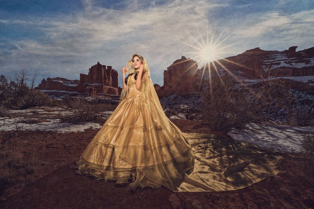 A golden ballgown dress flows dramatically against a desert landscape with red rock formations and sunburst in the background.