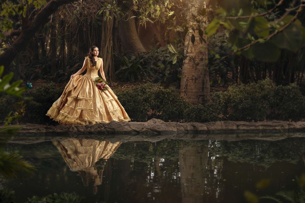 A golden vintage ball gown reflects in a tranquil pond surrounded by moody forest scenery at sunset.
