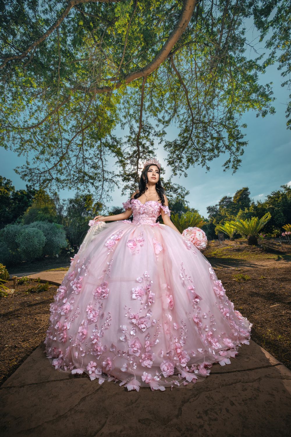A stunning pink ball gown with floral appliques photographed in a garden setting under hanging tree branches.