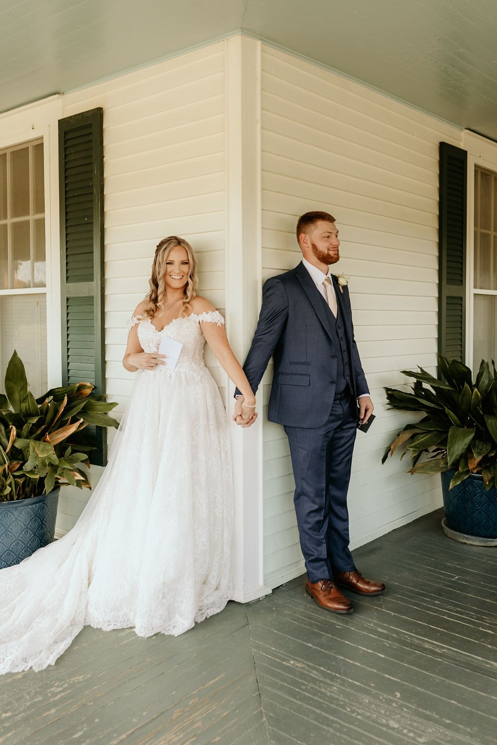 Photography of Couple stands apart during first look on porch of white farmhouse at wild daisy farm in Molena, Georgia