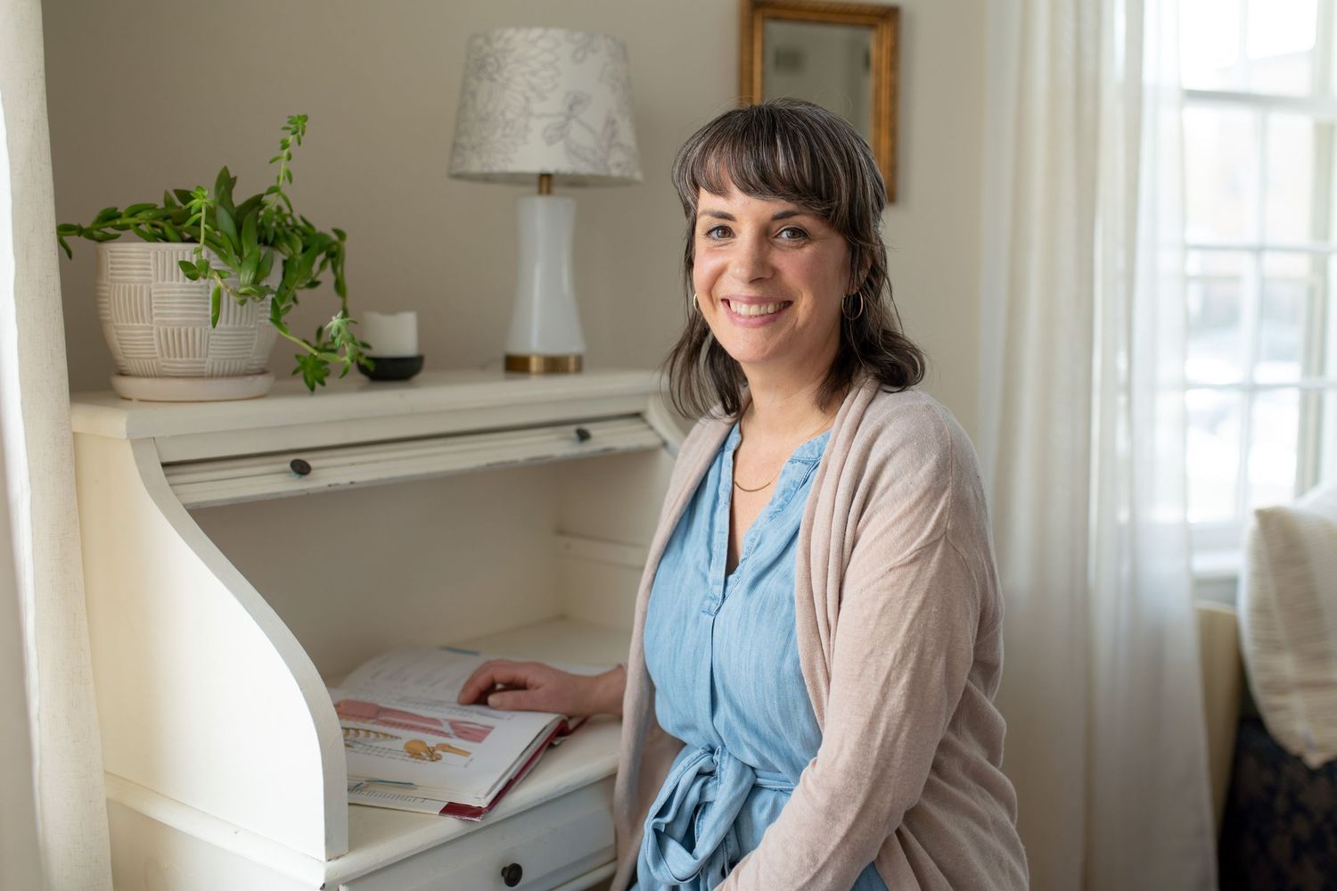 Someone sits at a white desk near a window in a bright room with a houseplant and table lamp visible.