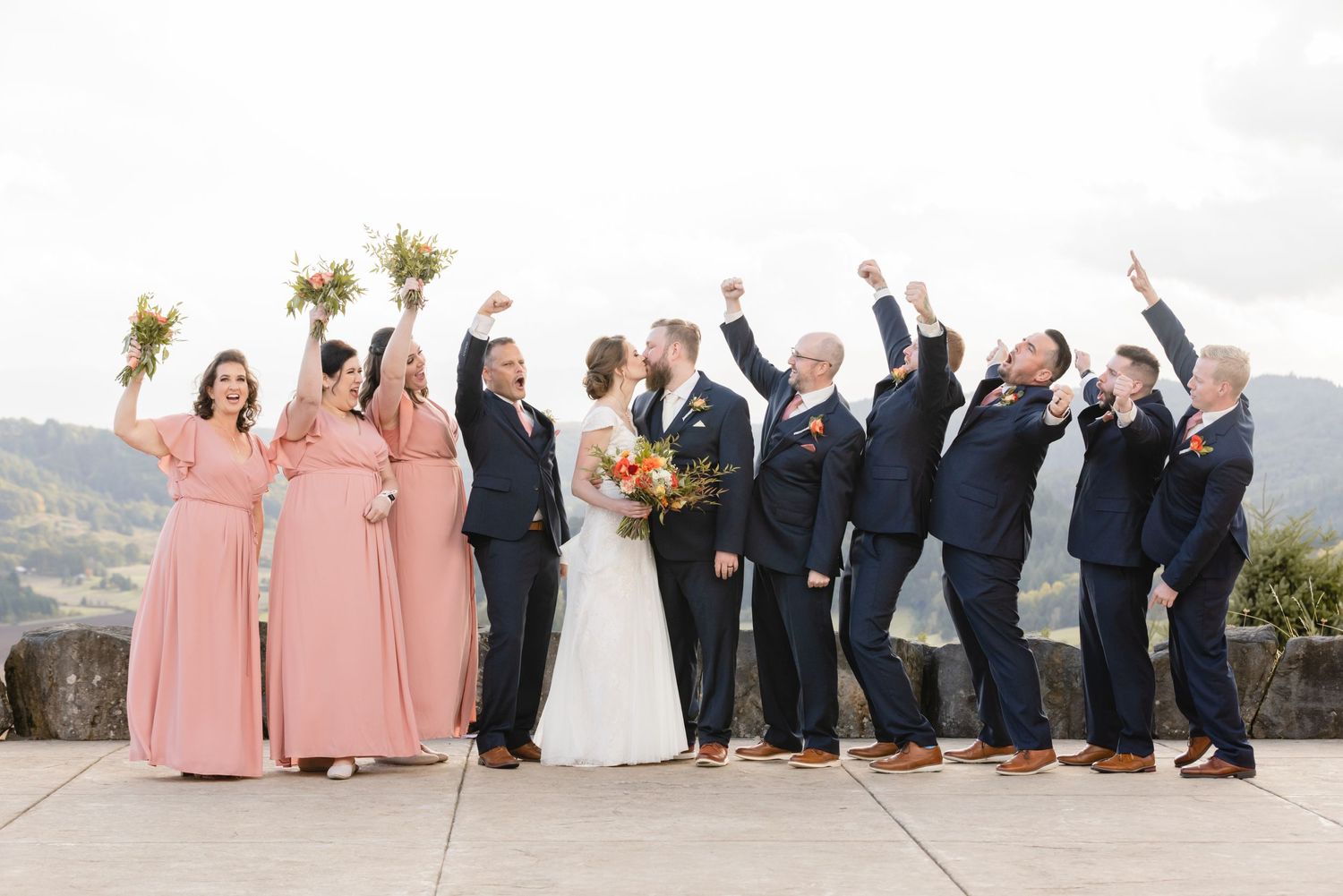Wedding party in pink bridesmaid dresses and navy suits celebrate joyfully against a mountain vista.