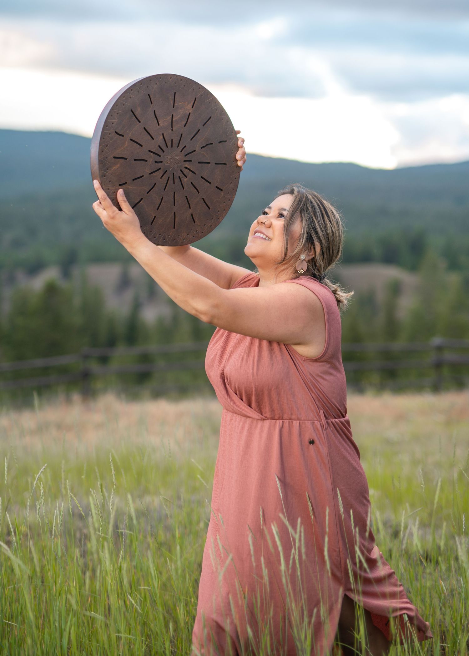 Indigenous woman in pink dress holding ocean drum overhead while standing in tall grass - Okanagan portrait by Tania Veitch.