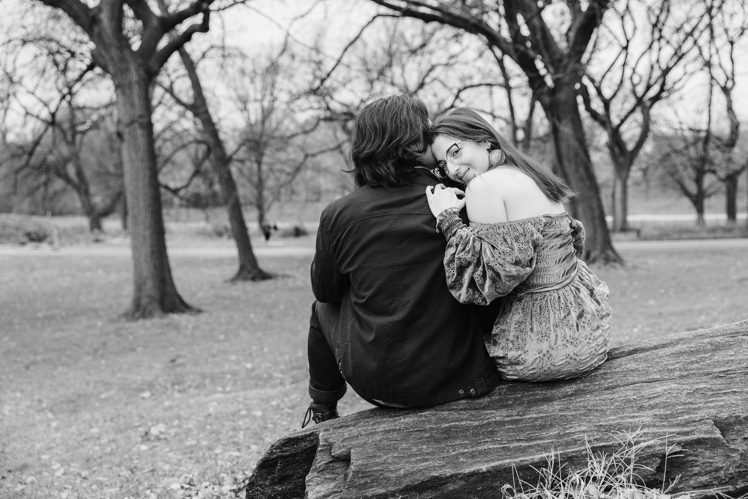 Black and white photo of couple sitting together on fallen tree in park.