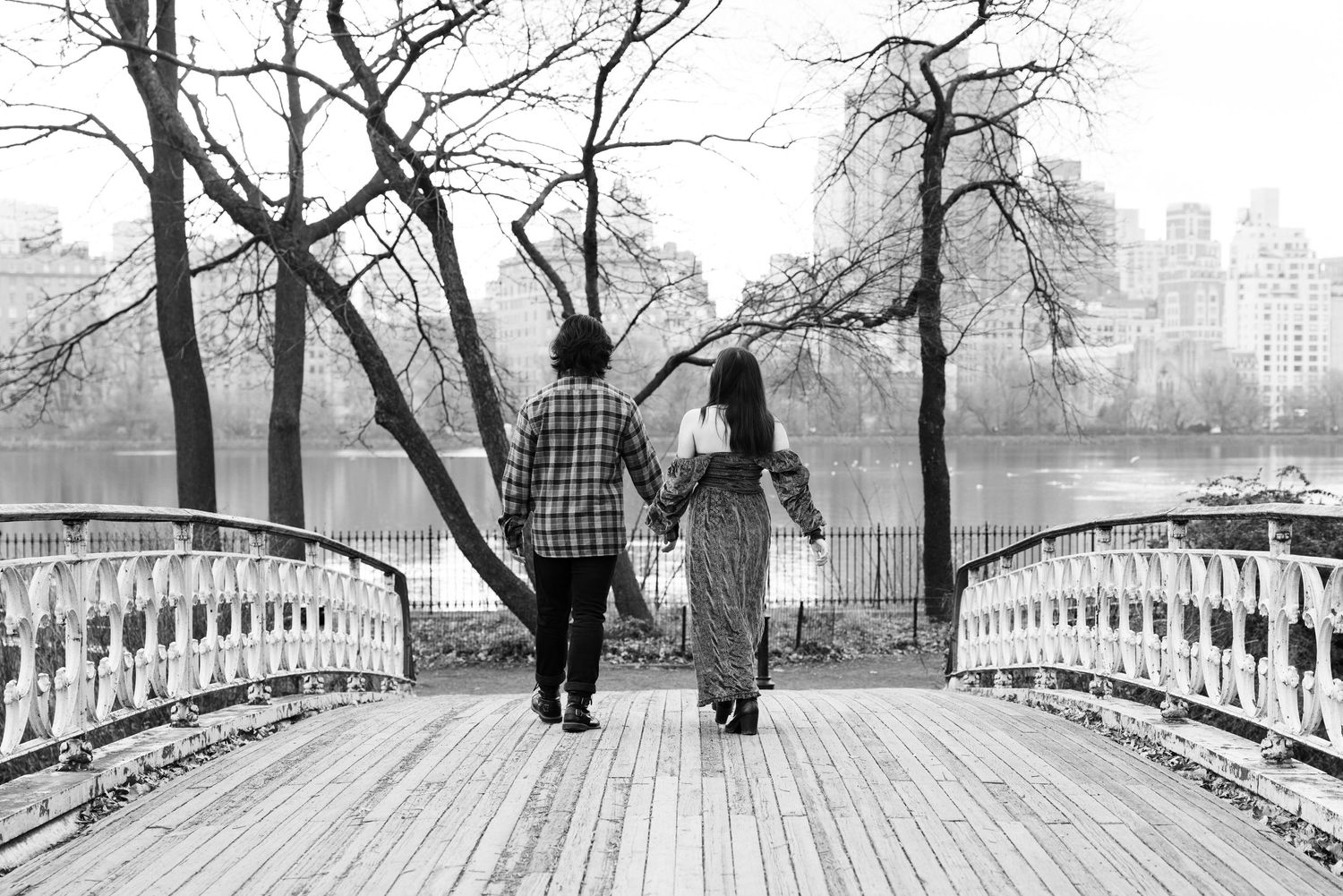 Black and white photo of couple walking across bridge in city park.