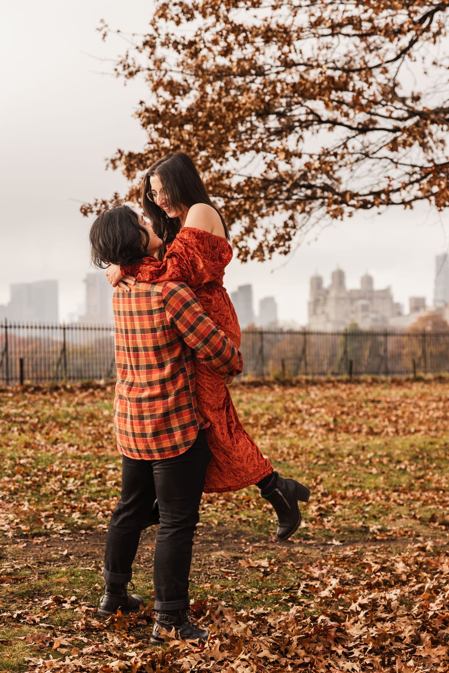 A couple embraces in a playful lift against an autumn cityscape background.