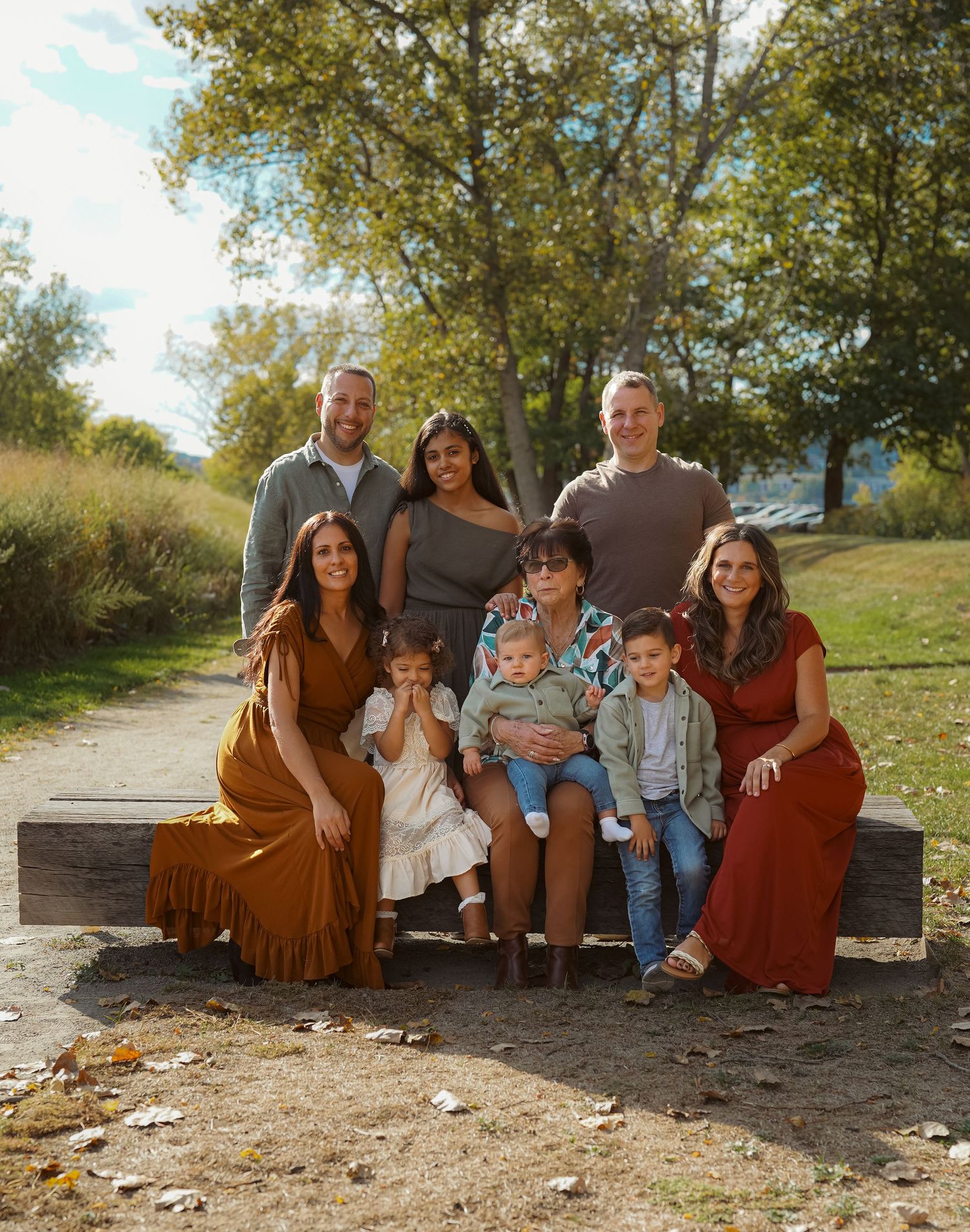 A family sits together outdoors on a wooden bench surrounded by autumn trees and fallen leaves.