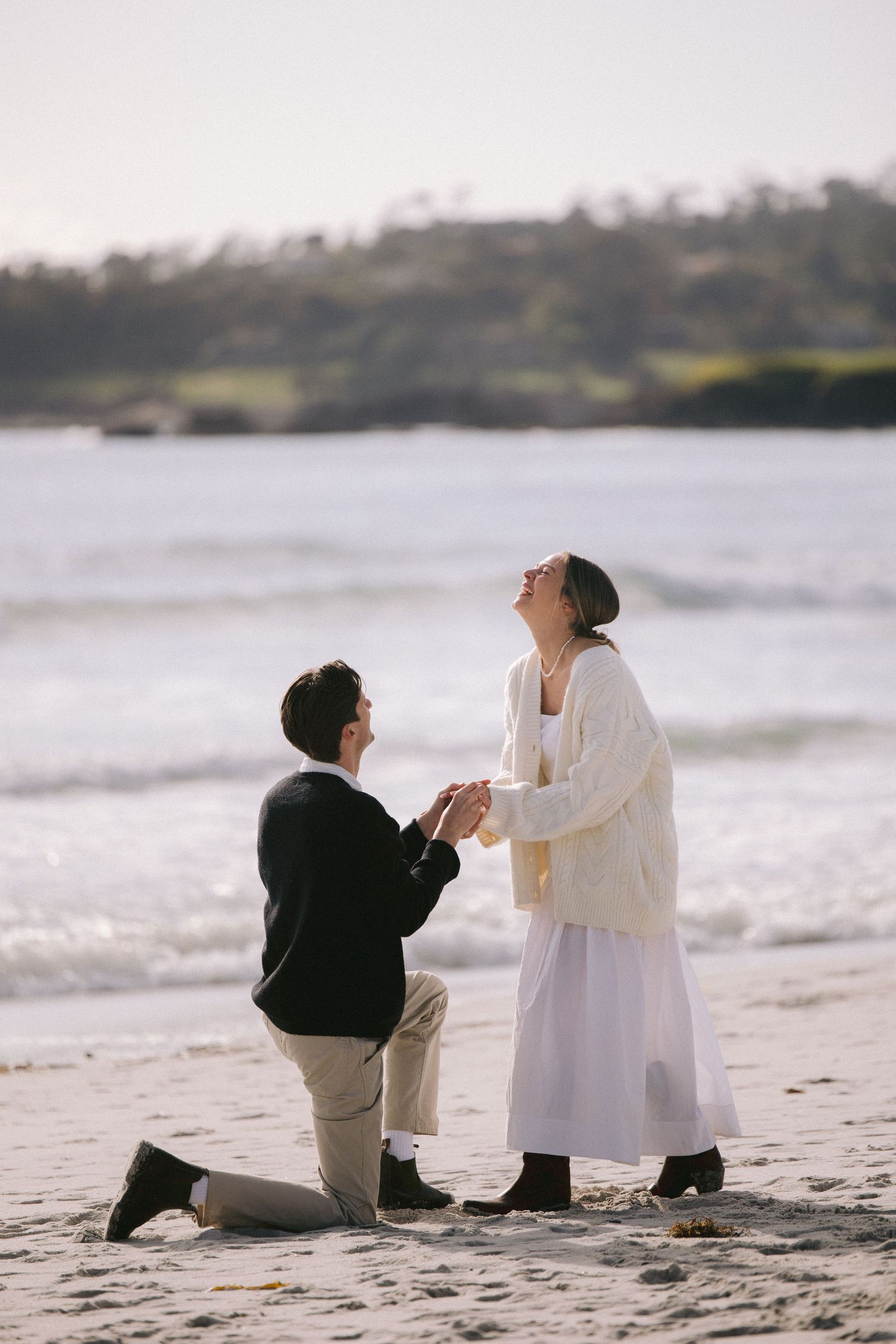 A couple shares a romantic proposal moment on a beach with crashing waves and coastal scenery in the background.