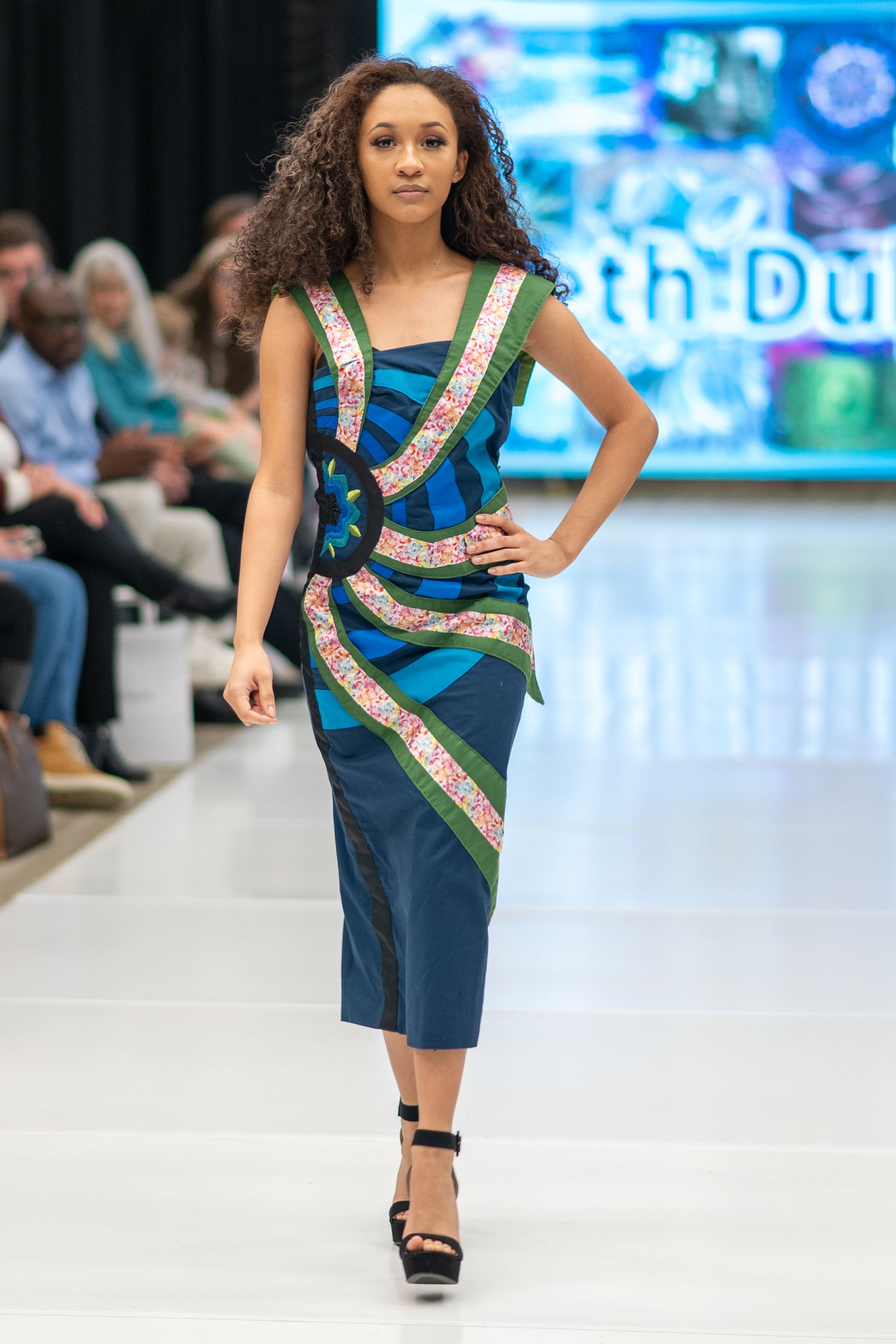 Model walks runway in blue fitted dress with decorative metallic striped details and ankle-strap platform heels.