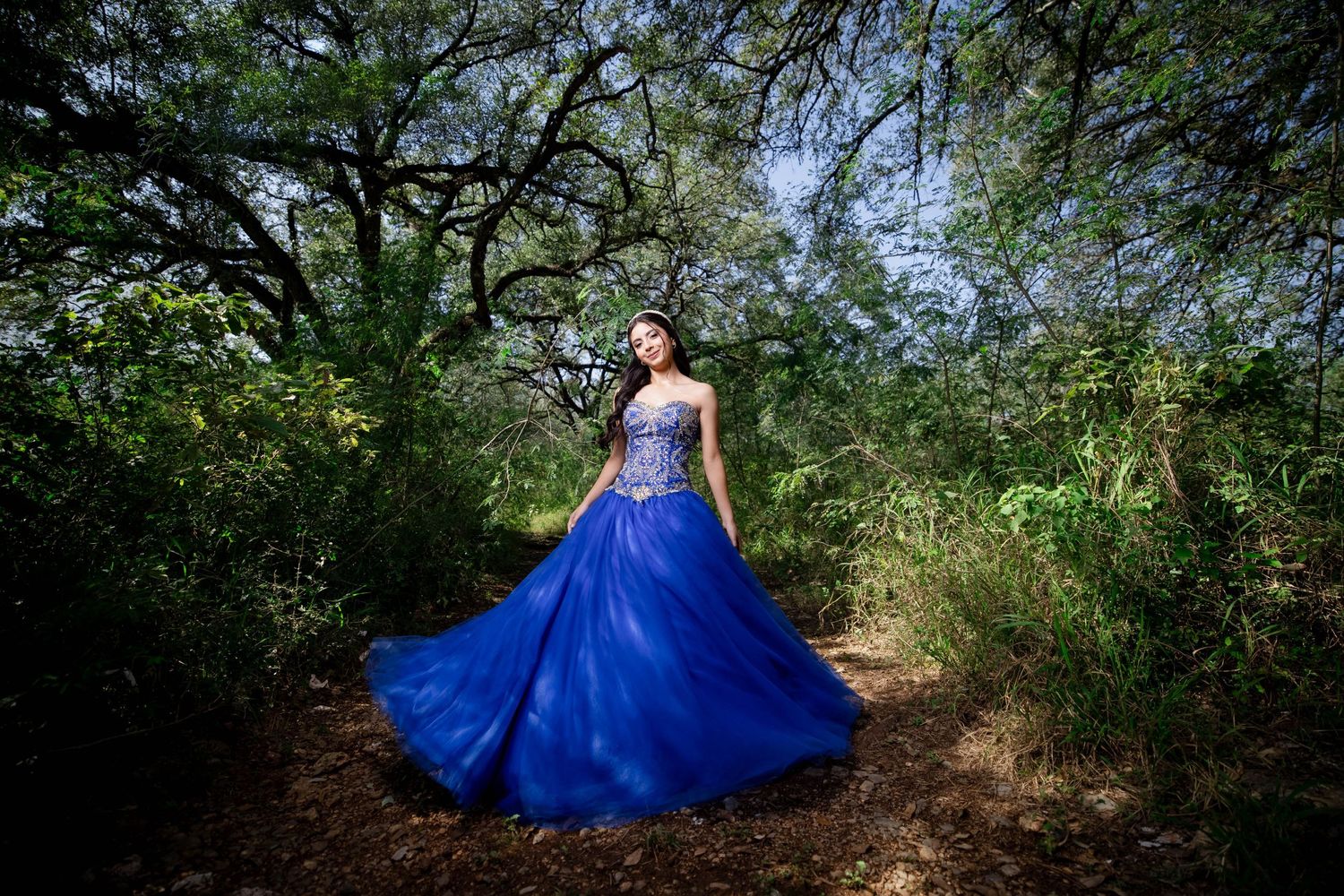 Stunning blue ballgown twirls on forest path surrounded by green foliage.