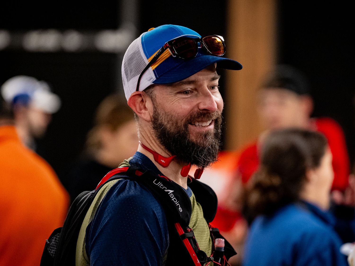 Trail runner wearing blue sunglasses and a white cap smiles during an outdoor race event.