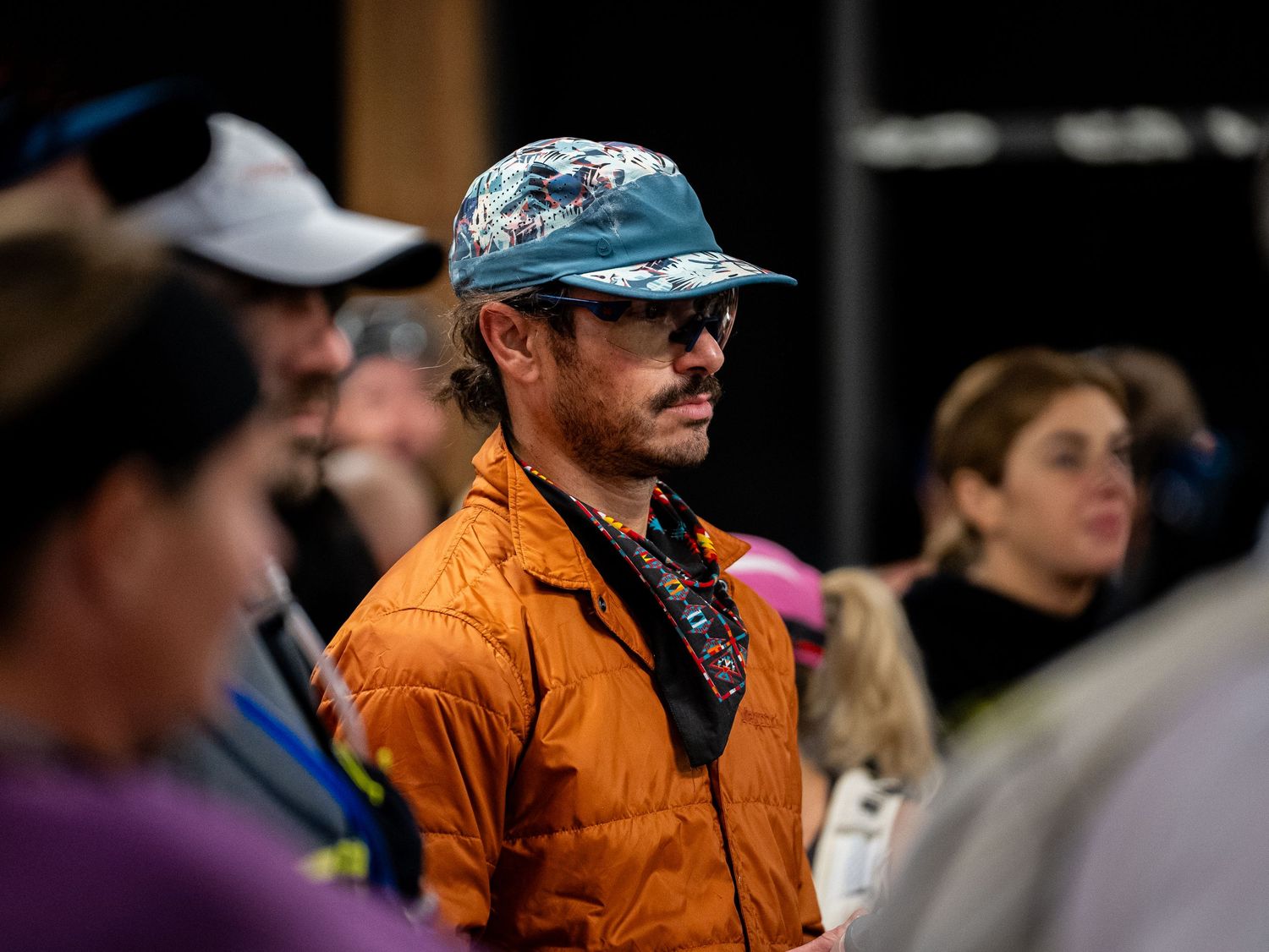 Runner in orange jacket and camo hat stands among crowd at outdoor endurance event.