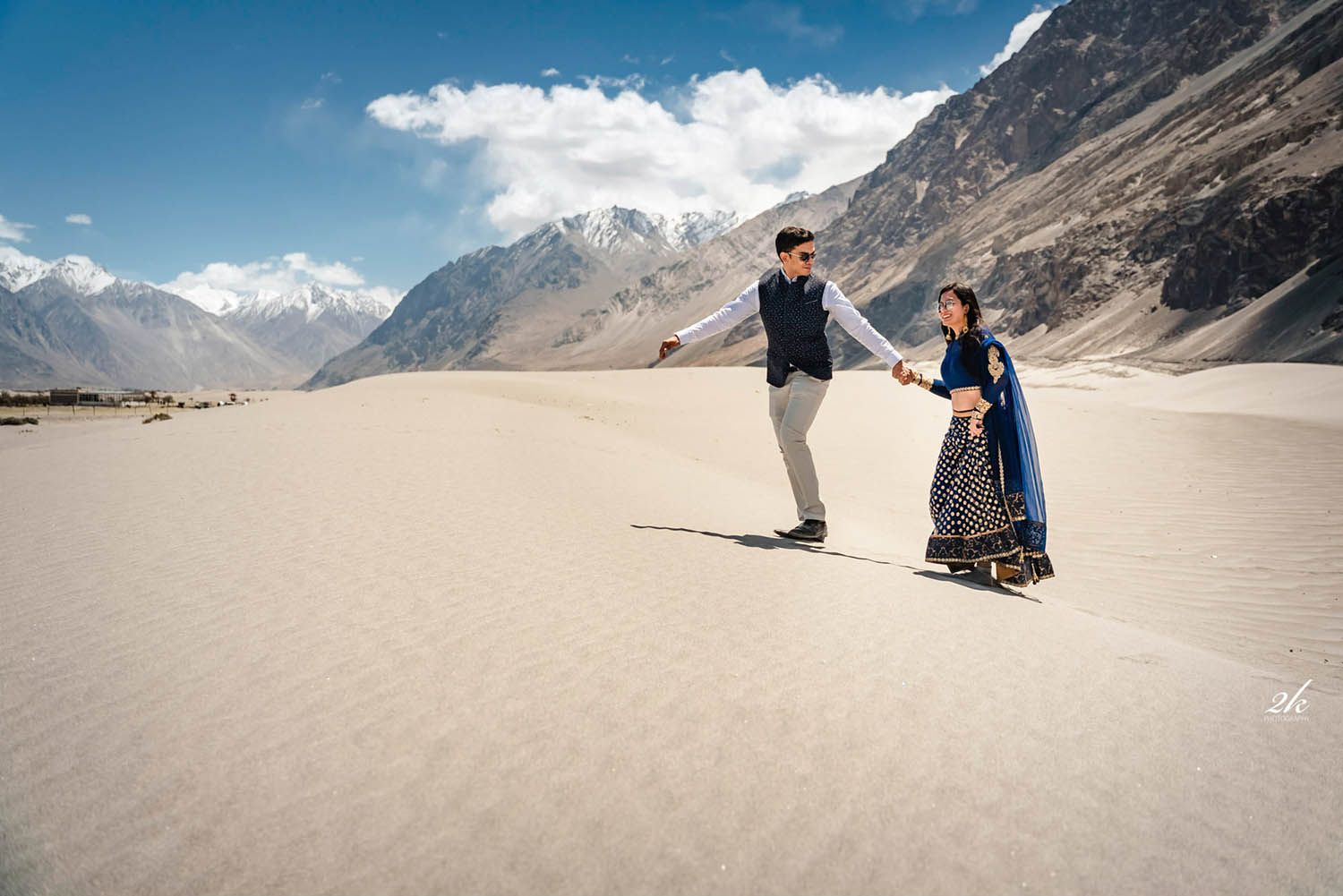 Two people holding hands in a traditional outfit stand on sandy terrain surrounded by dramatic mountain peaks.