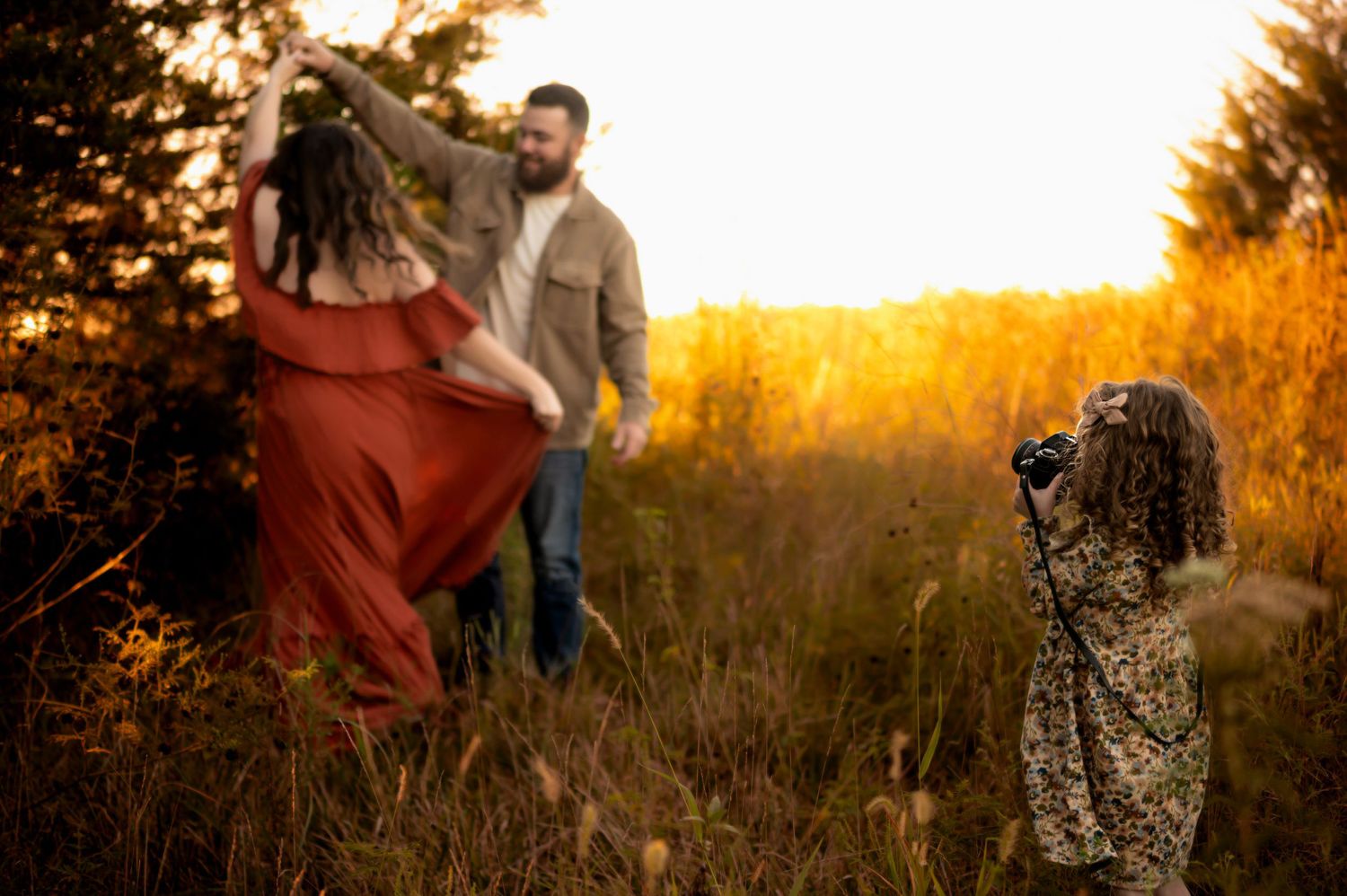 A couple dances together in a golden field at sunset while a camera on tripod captures their moment.