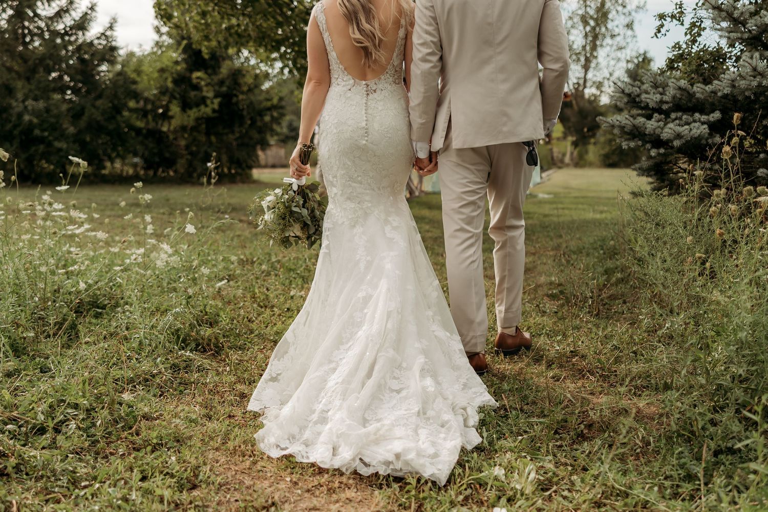 Wedding party in sage green dresses and tan suits gather outdoors under rustic wooden pavilion on sunny day.
