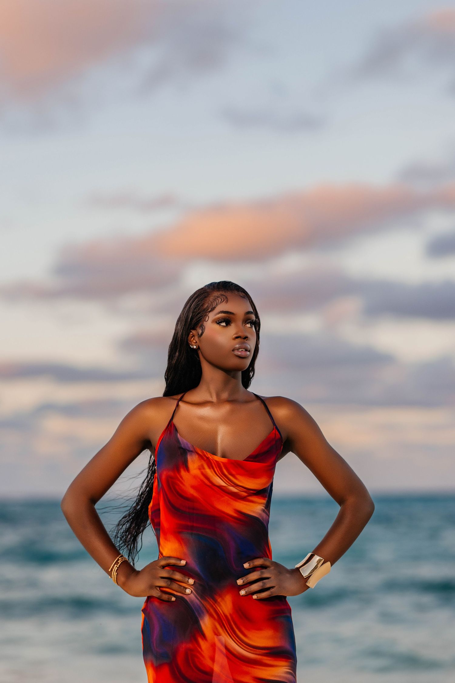 Model stands confidently in colorful dress against ocean sunset backdrop.