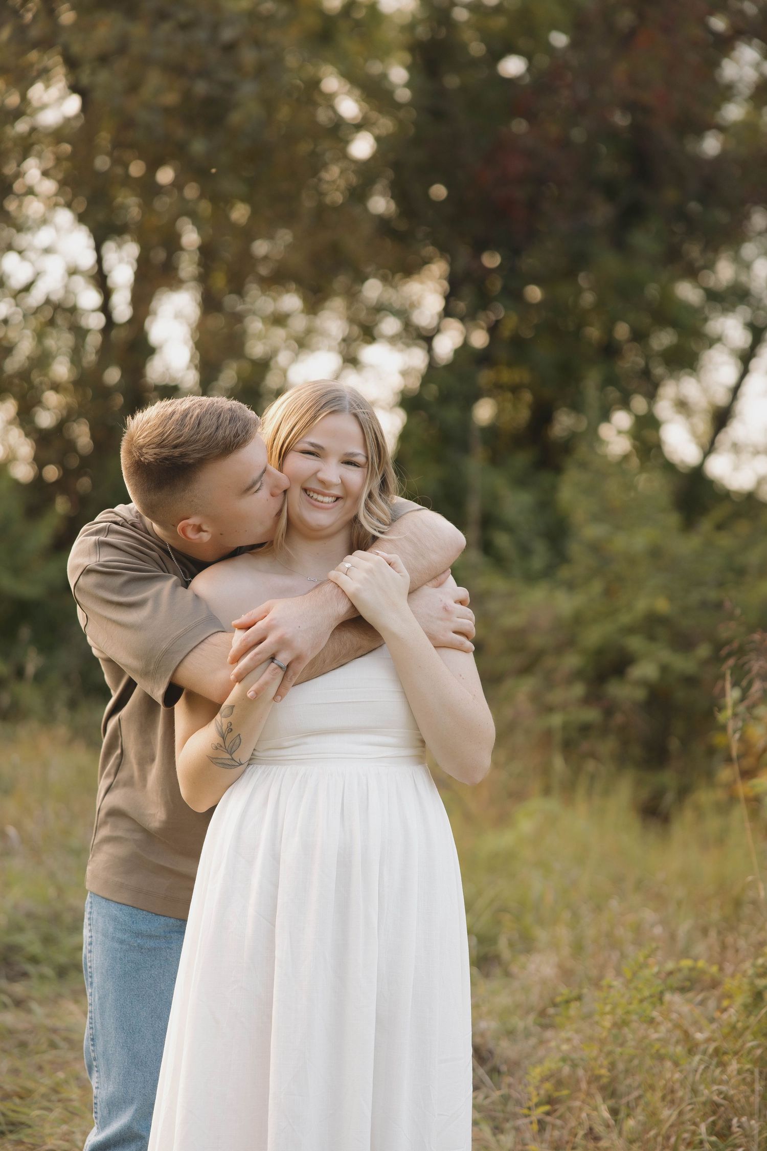 A loving moment captured outdoors as couple embraces in a sunlit field with trees in the background.