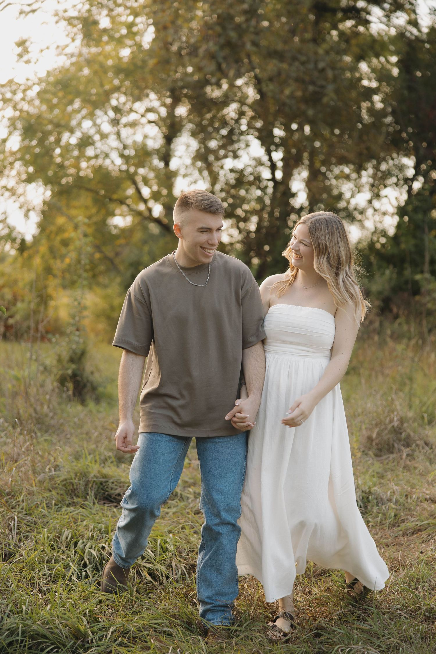 A couple walks hand in hand through a sunlit field, with her in a flowing white dress and him in casual attire.