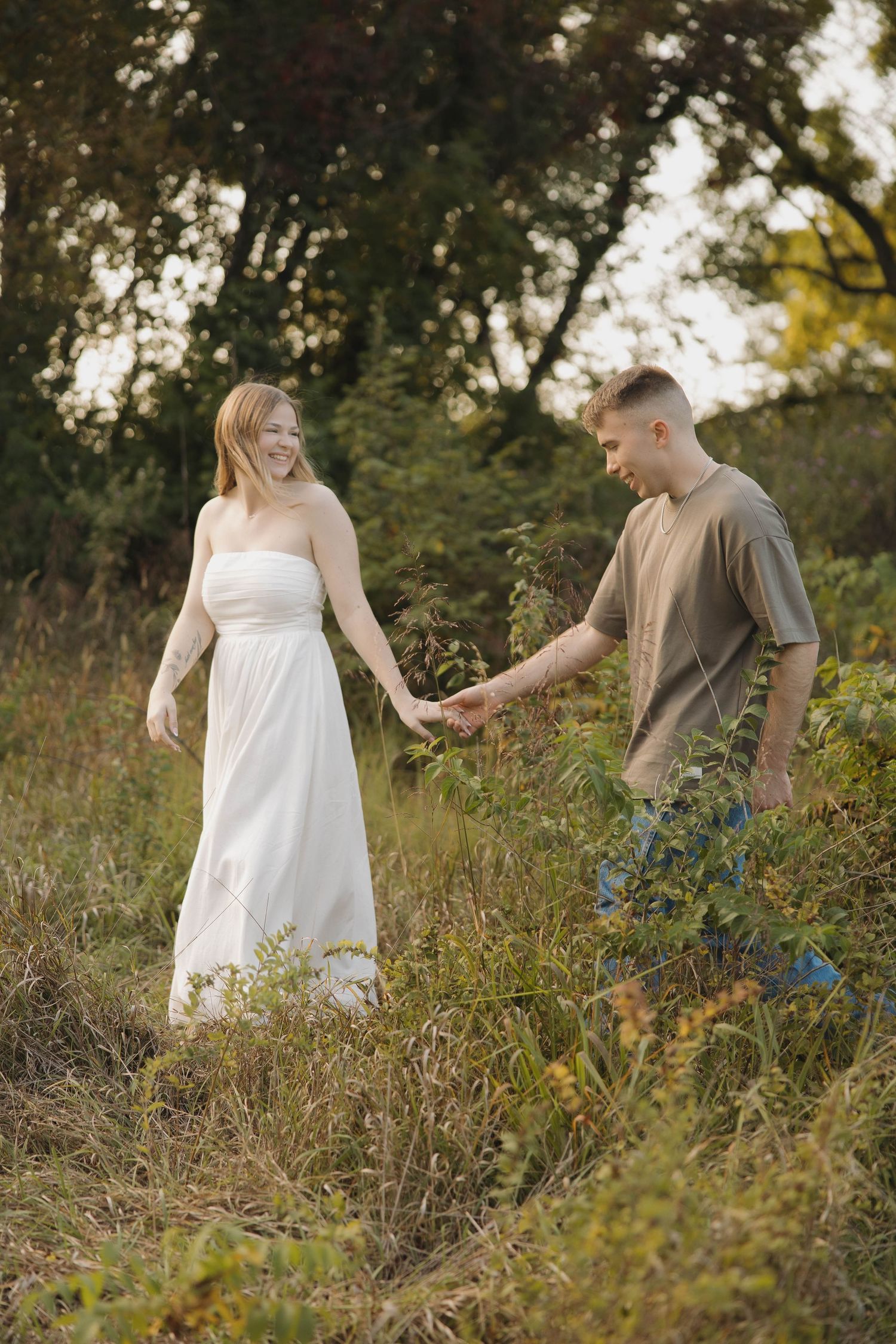 A couple walks through a sunlit wildflower meadow with tall grass and natural vegetation during golden hour.