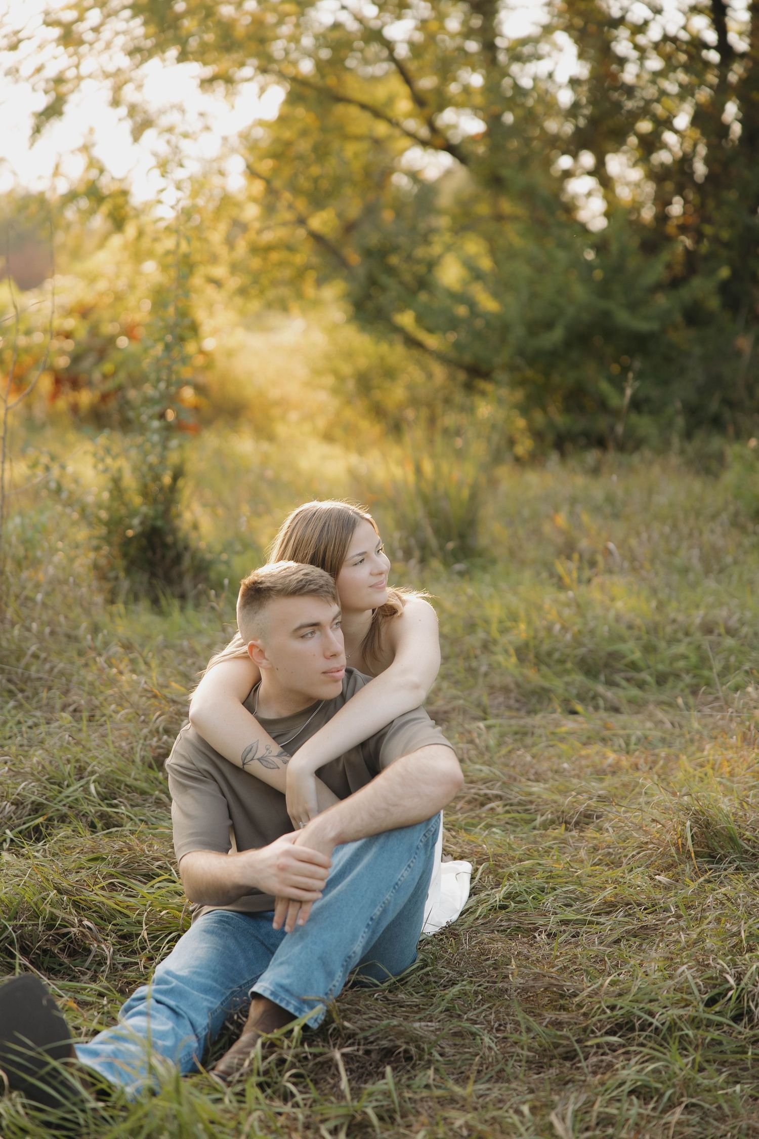 A romantic couple sits together in tall grass during golden hour in a natural outdoor setting.