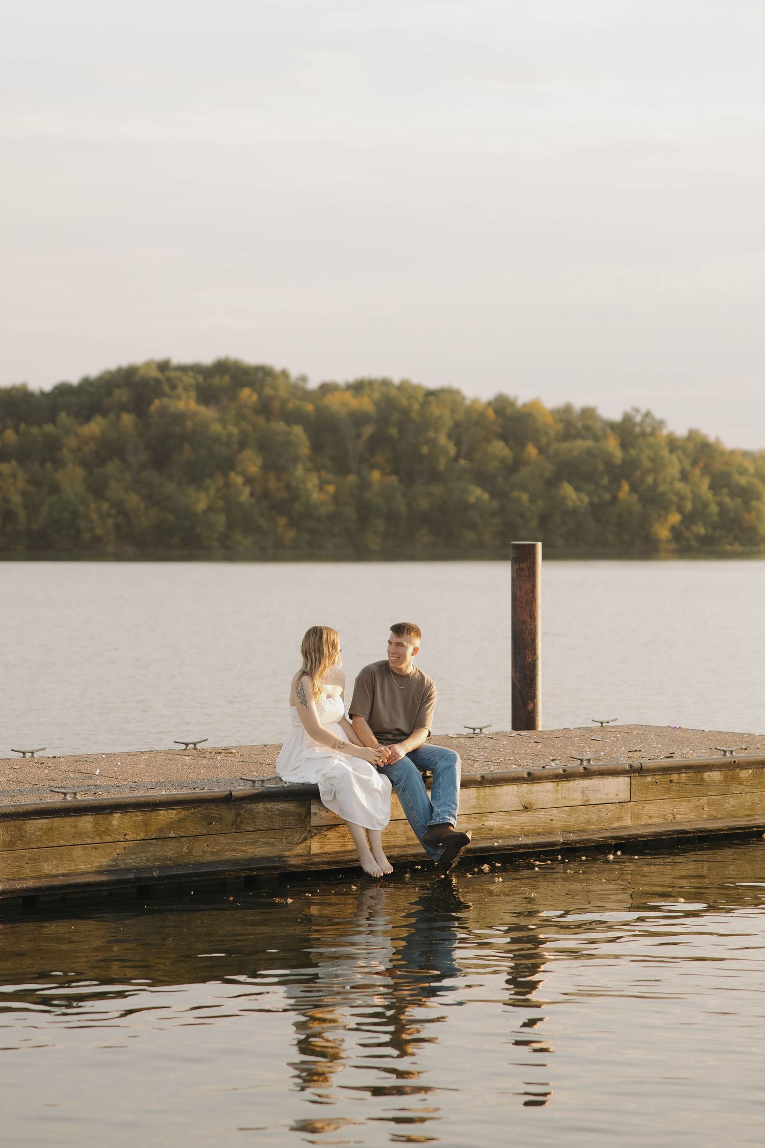 A romantic sunset moment of a couple sitting on a wooden dock by the lake with autumn trees in the background.