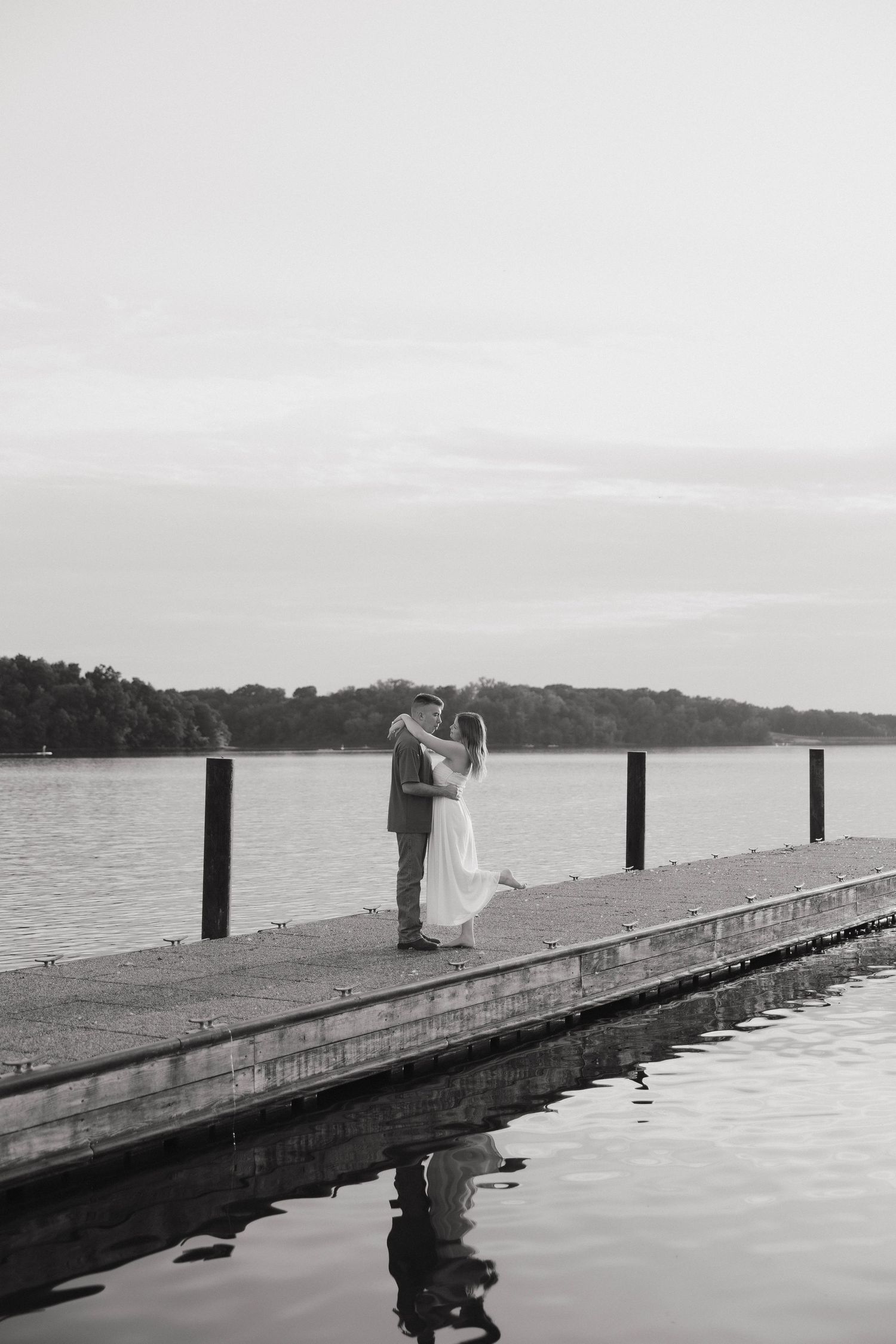 A romantic couple embraces on a wooden dock overlooking a serene lake at sunset in black and white.