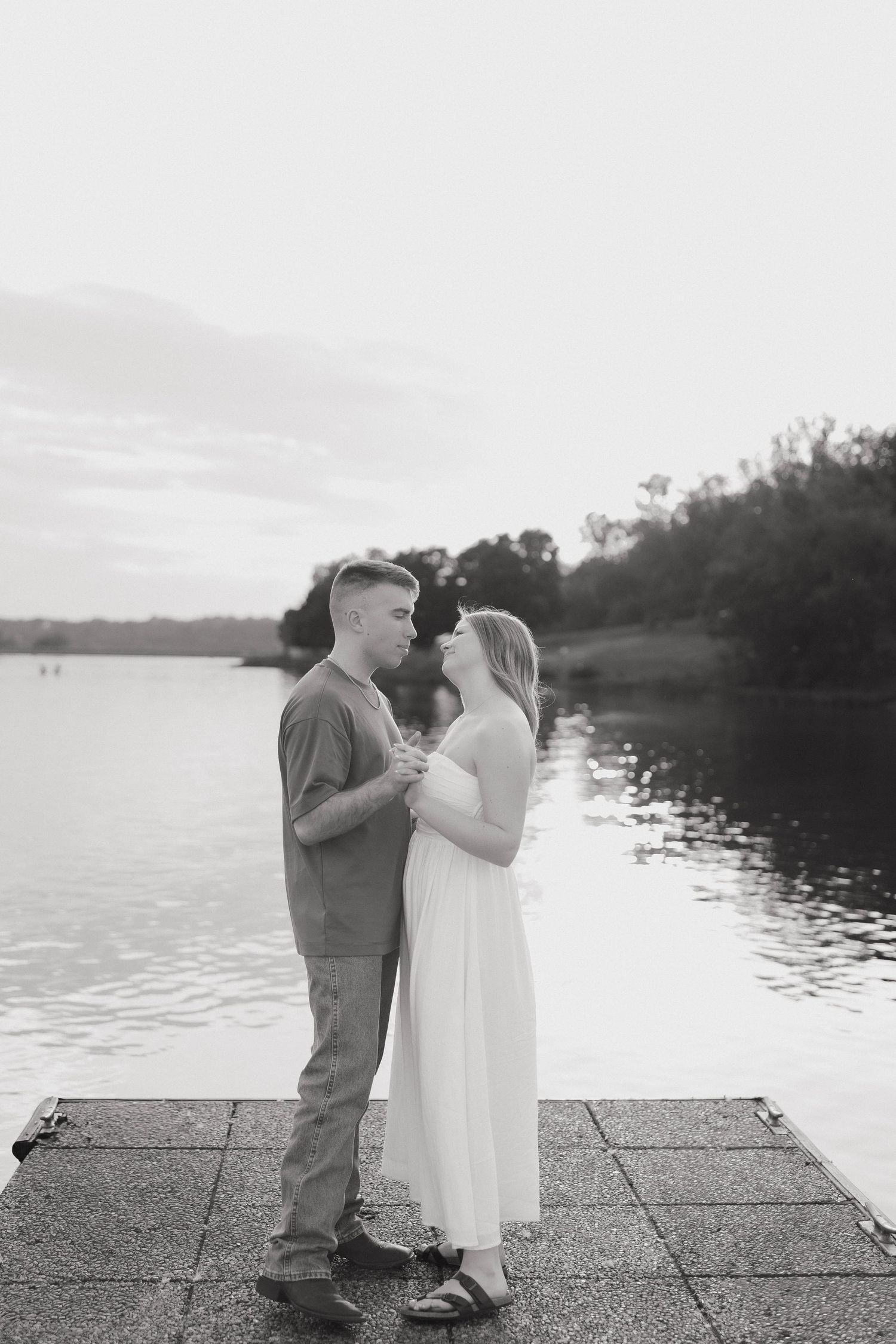 A couple shares an intimate moment on a dock by a lake during sunset in black and white.