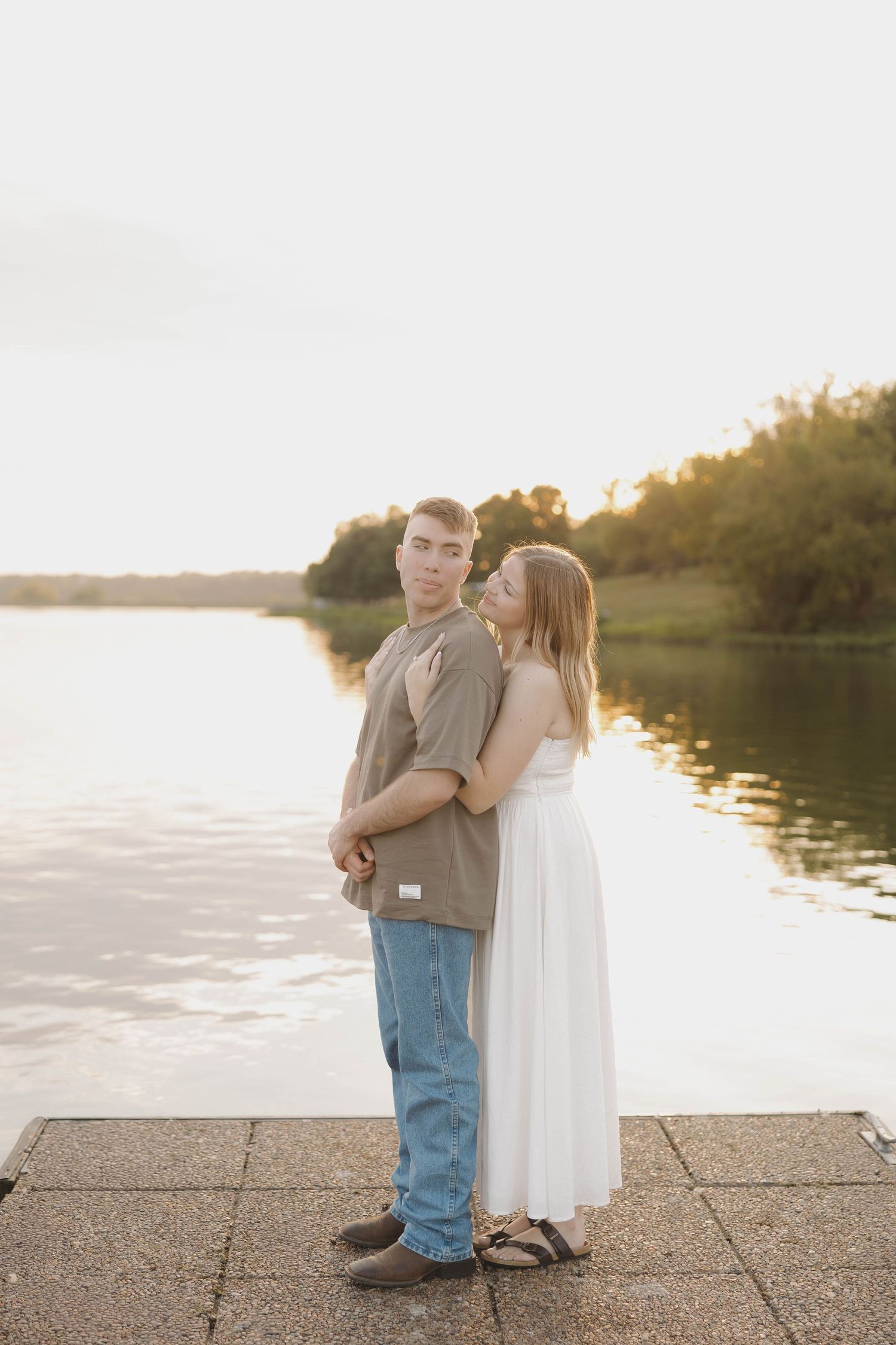 A couple embraces on a concrete pier by a lake during golden hour sunset.