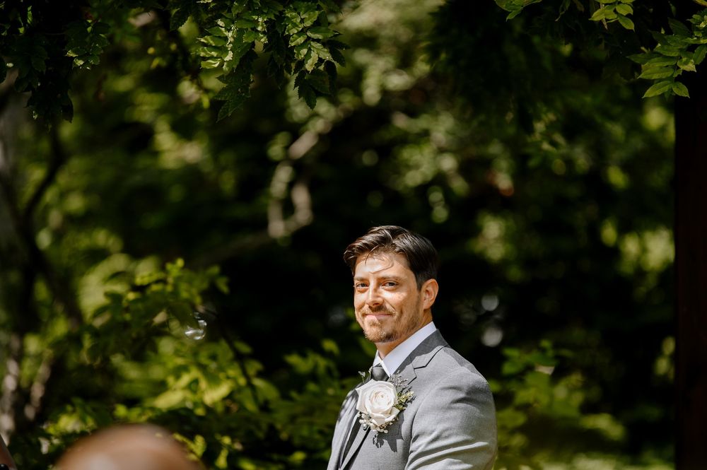 A person in a grey suit stands outdoors amidst lush greenery and forest foliage on a sunny day.