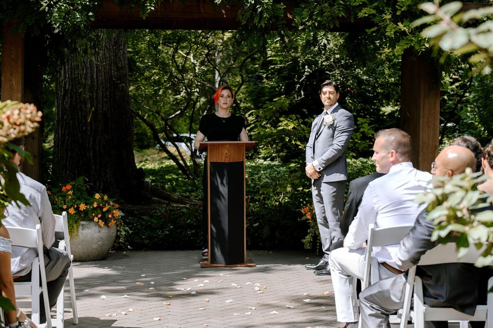 An outdoor wedding ceremony takes place in a garden setting with guests seated and officiant at podium.