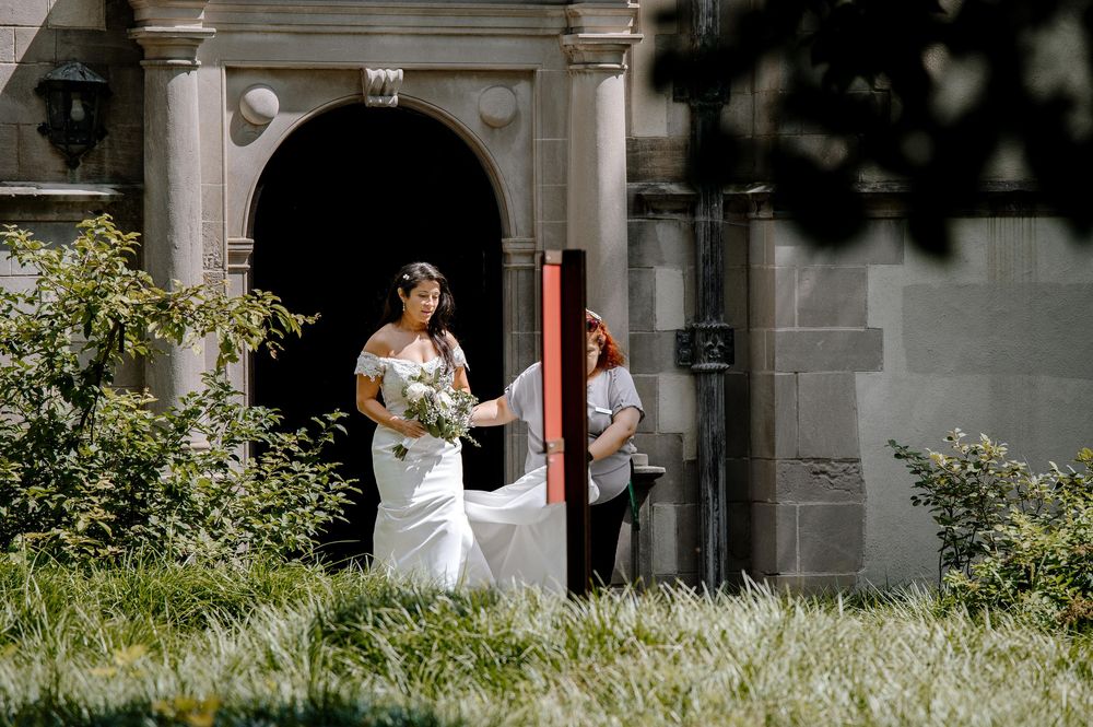A wedding couple stands near an elegant arched entrance surrounded by lush greenery on a sunny day.