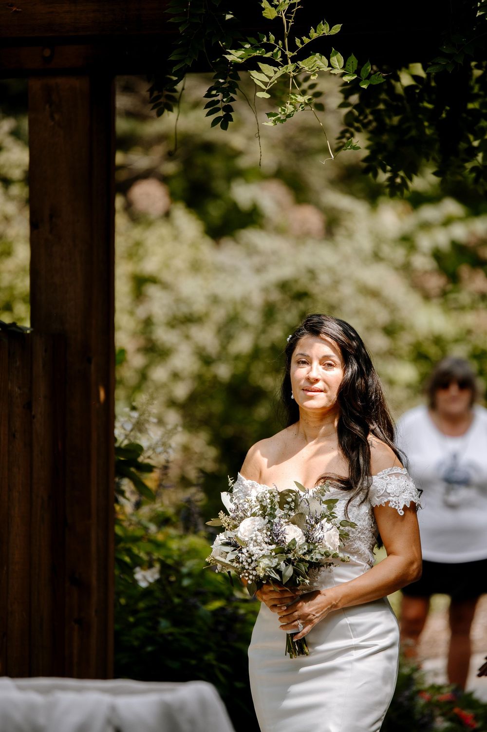 A bride in an off-shoulder white wedding dress holds a bouquet while standing in a garden setting.