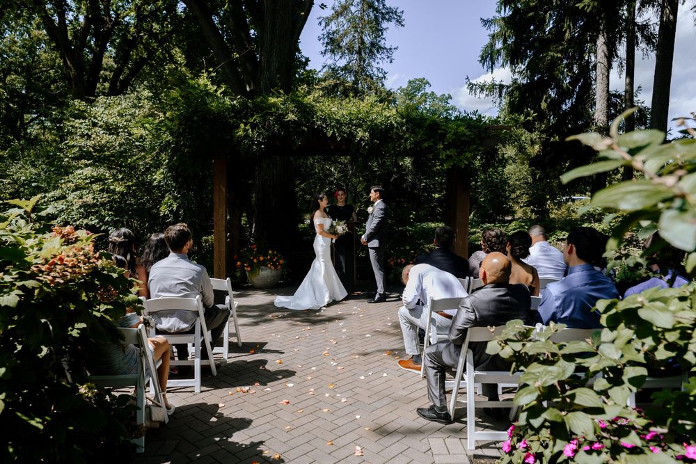Guests gather outdoors at a sunny garden wedding ceremony featuring rows of white chairs and natural landscaping.