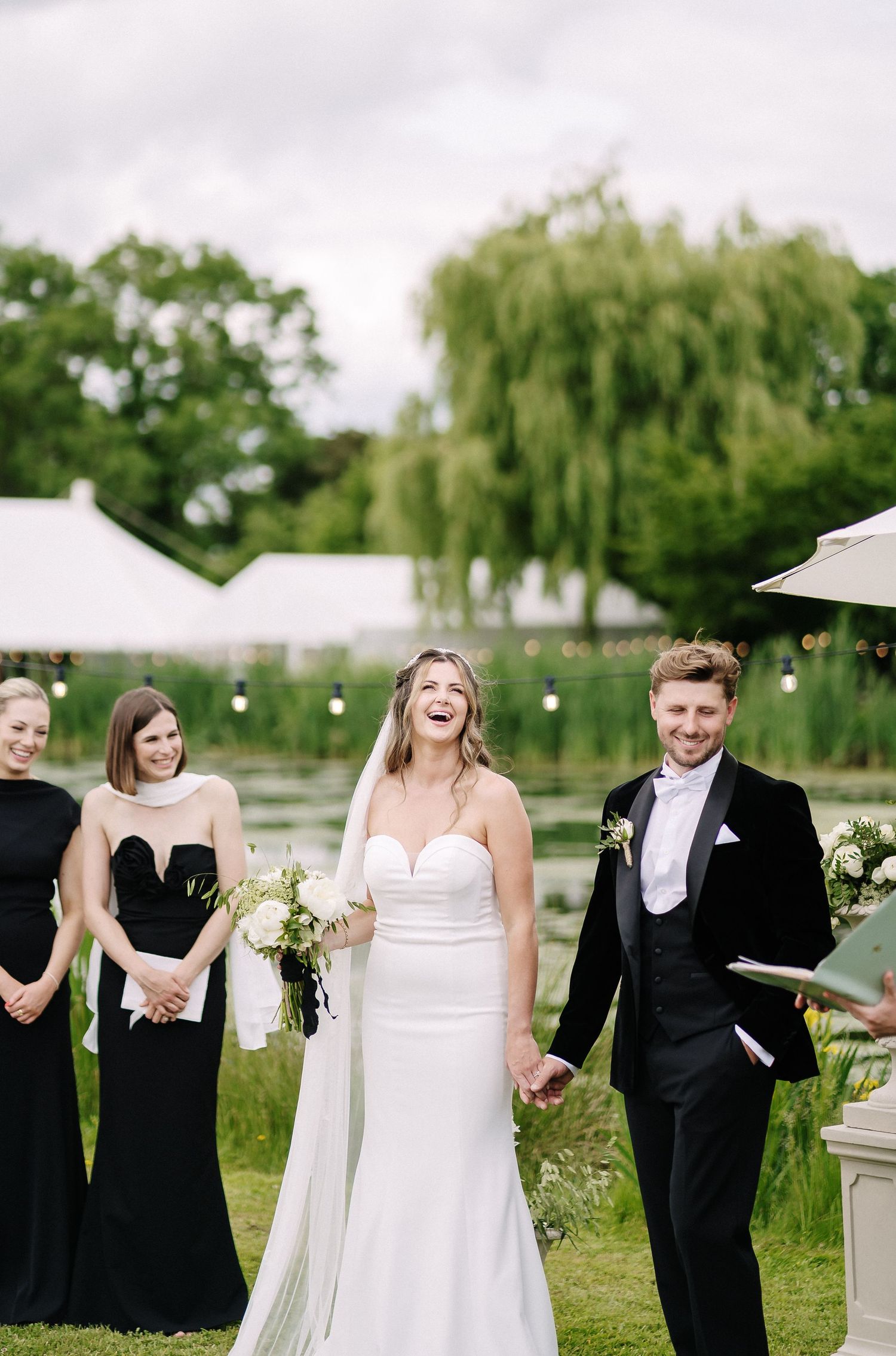 A wedding party in formal black and white attire walk together and laugh outdoors on grass with willow trees behind them.