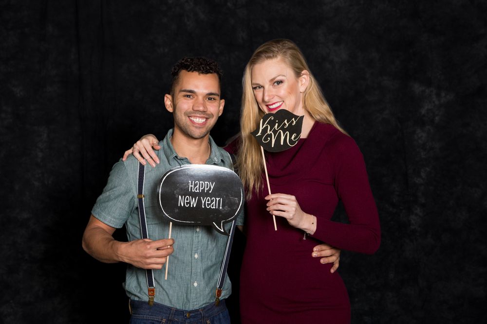 Couple posing at New Year photo booth, man holding Happy New Year sign, woman holding Kiss Me prop, dark background.