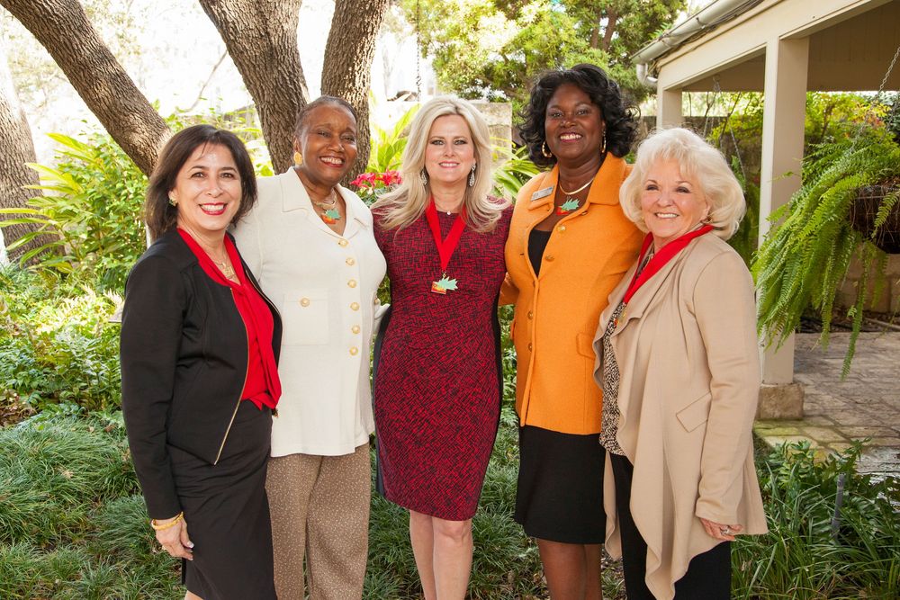 Five diverse women smiling and posing together outdoors in a garden setting, dressed in professional attire.