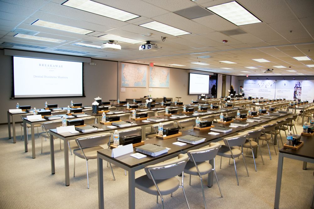 Modern computer lab classroom with rows of desks, monitors, and a projector screen in a bright, well-lit corporate training room.