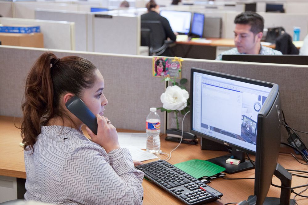 Woman talking on phone at office desk with computer, keyboard, and coworker visible in open plan workspace.