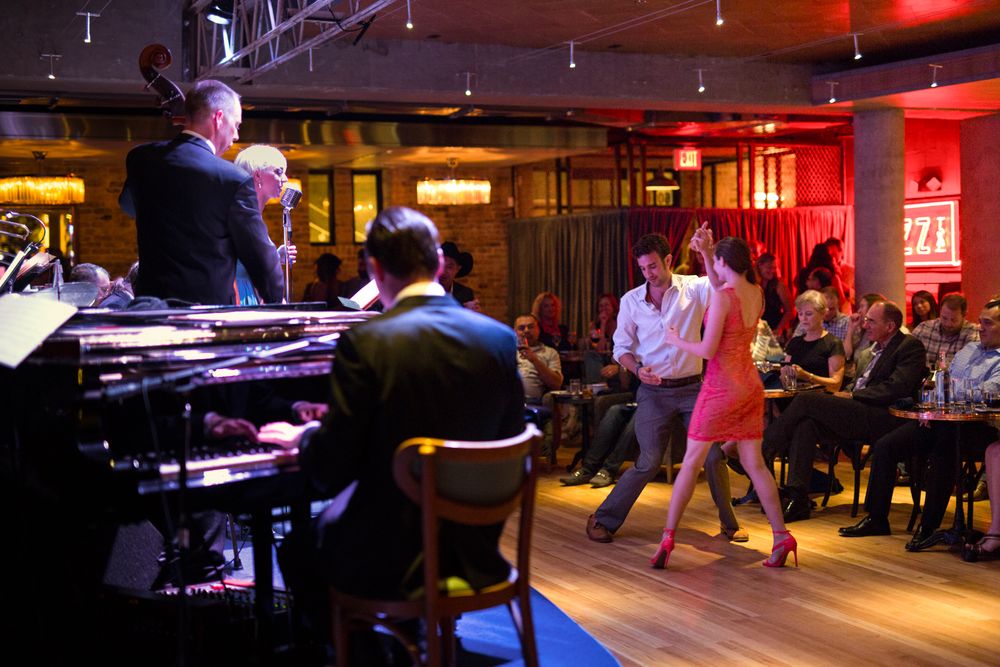 Live jazz band performs as couple dances on wooden floor in elegant red-lit venue with seated audience.