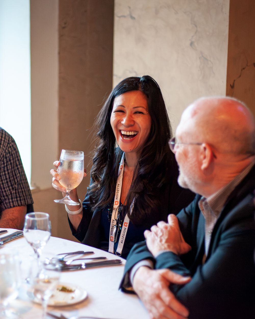 Woman laughing at a dinner table holding a wine glass, seated next to a man, both enjoying a social event.